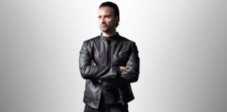 Modern Field Leather Jacket: Fresh Urban Look with Function Man wearing a fitted black leather field jacket with zippered pockets, standing with arms crossed against a light gray gradient background."