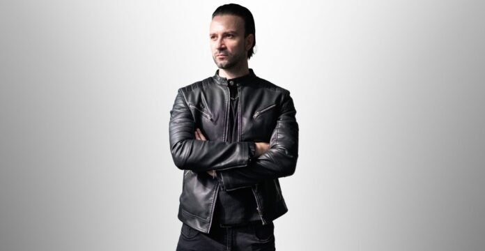 Man wearing a fitted black leather field jacket with zippered pockets, standing with arms crossed against a light gray gradient background.