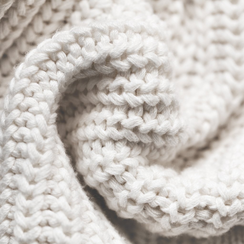 A close-up photograph of chunky cream-colored knitted fabric, showing thick interwoven stitches and a soft, cozy texture.