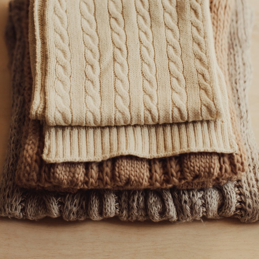 Stack of folded knitted sweaters in cream, beige, brown, and gray, with cable-knit and ribbed textures. Naturally Odor Resistant