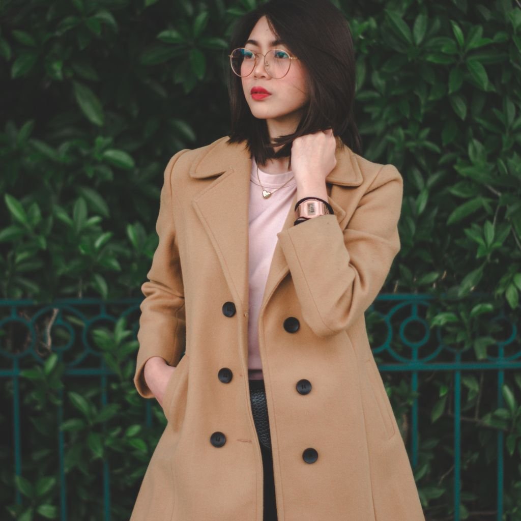 A stylish woman stands outdoors wearing a camel double-breasted coat over a light pink top, glasses, and a rose-gold watch, with green foliage in the background.The types of overcoat