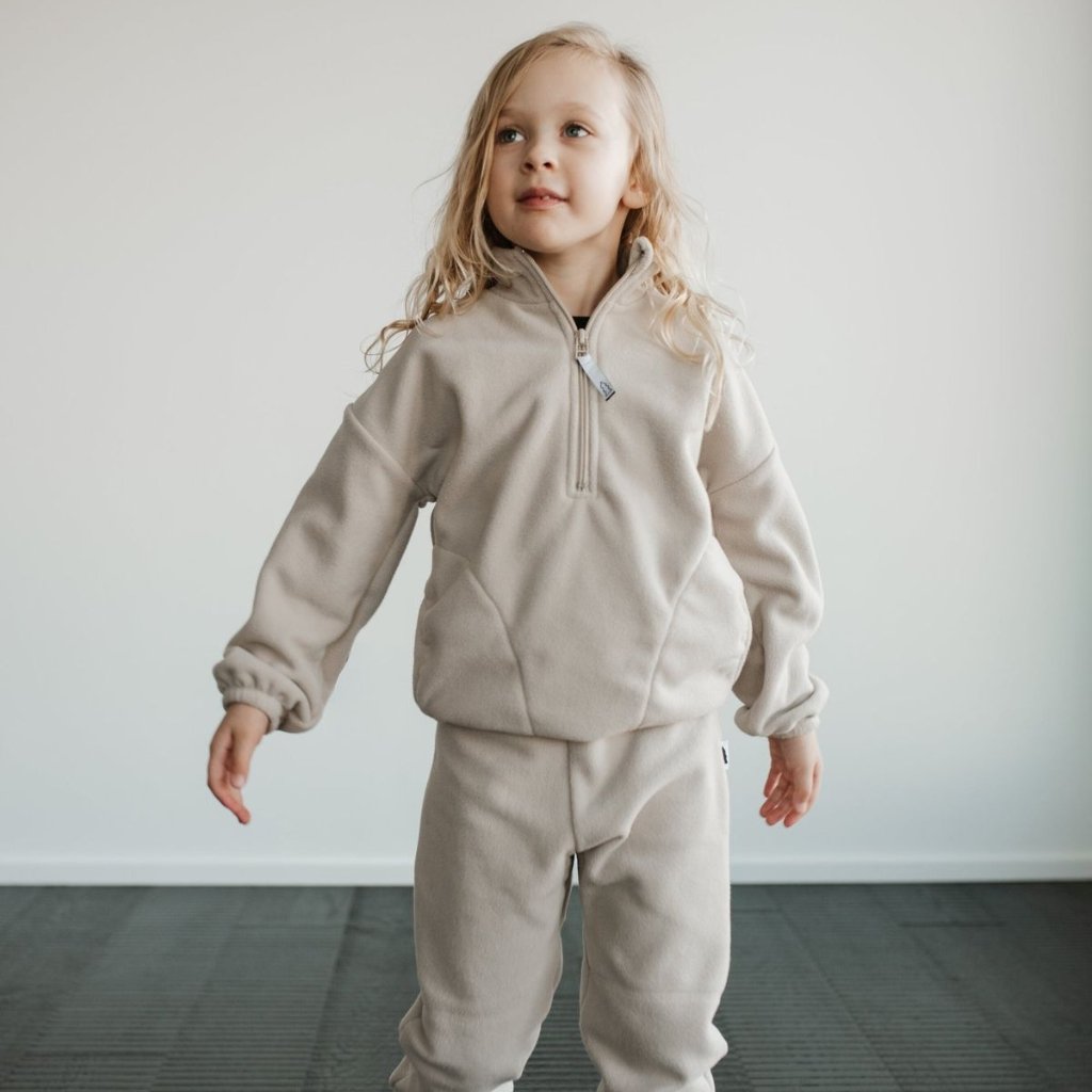 A young girl wearing a light beige fleece tracksuit stands indoors, looking slightly upward with a gentle smile.