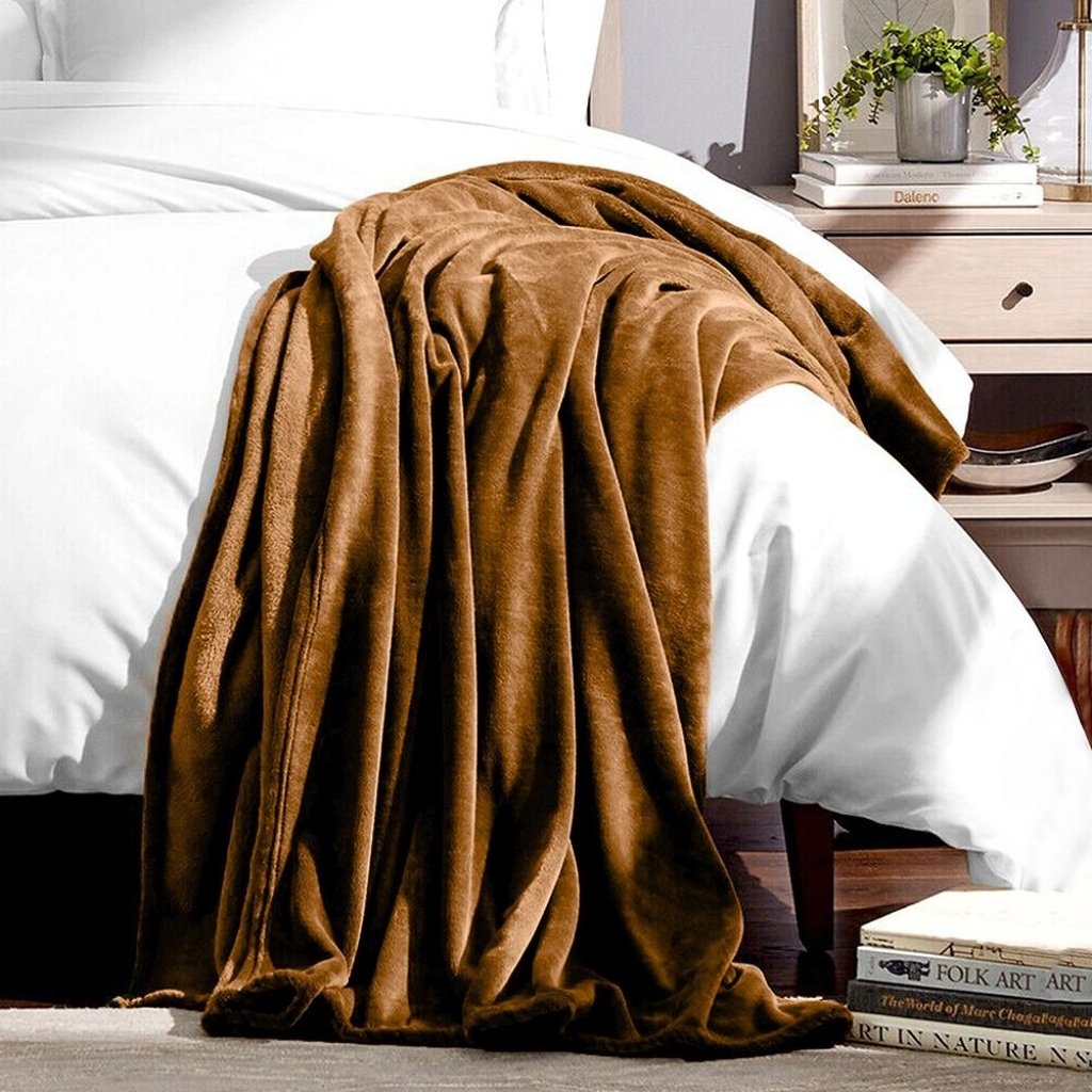 A cozy brown blanket draped over the side of a white bed in a neatly styled bedroom, with a nightstand and stacked books in the background.