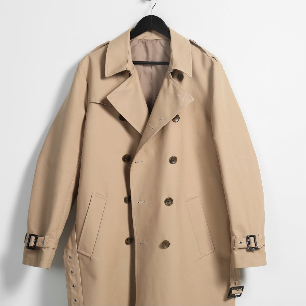 A beige double-breasted trench coat on a hanger, showing the front view with wide lapels, belt loops, and side pockets.

