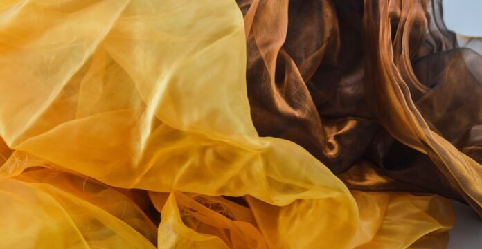 A close-up view of sheer silk organza fabric in warm amber and honey tones, with delicate, translucent folds and soft light.
