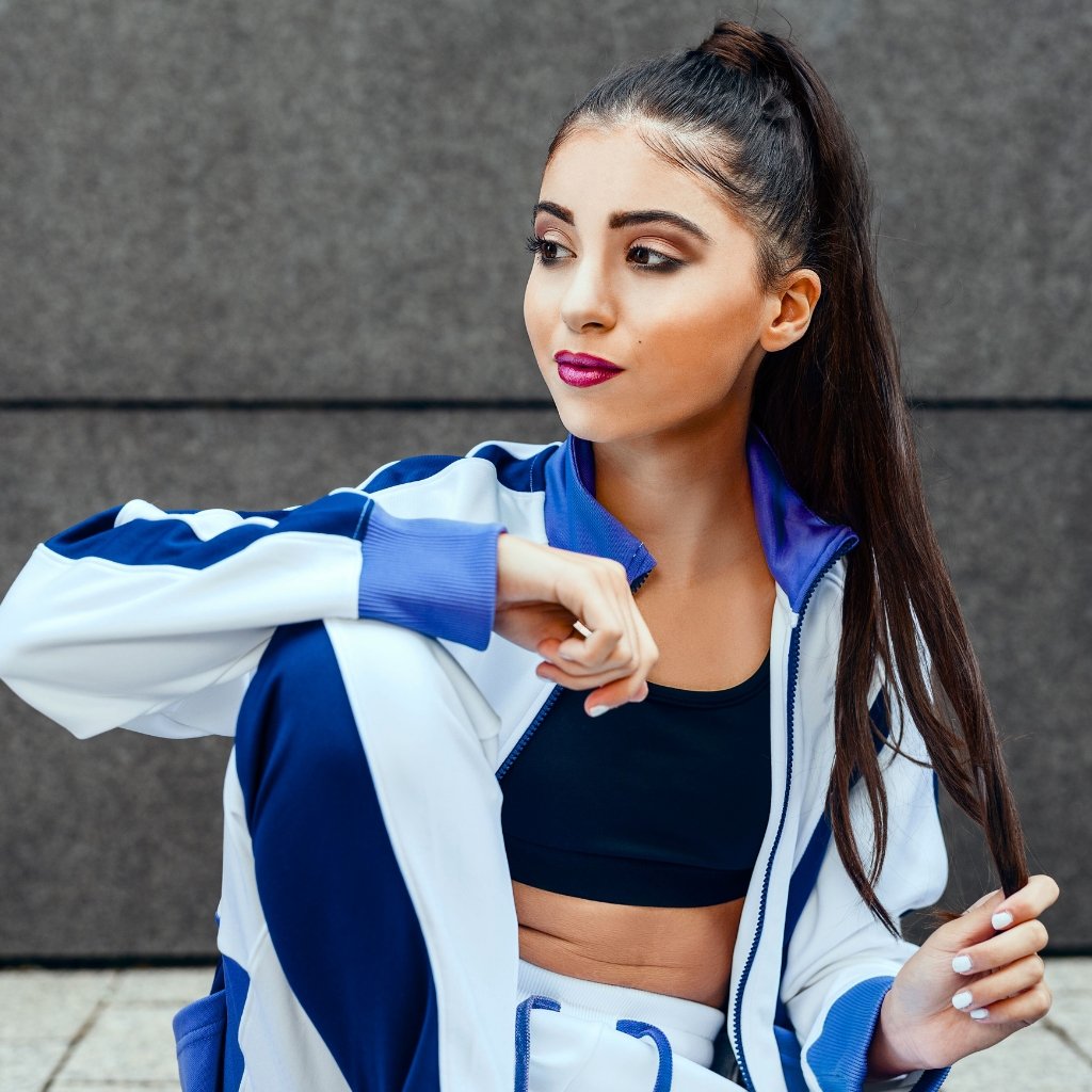 A stylish young woman with a high ponytail wearing a navy sports bra and a white-and-navy tracksuit jacket, looking to the side while touching her sleeve.