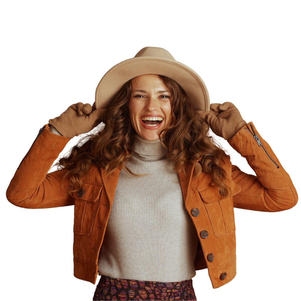 A smiling woman wearing a tan wide-brim hat, a tan/taupe turtleneck sweater, and an orange-brown suede jacket. She holds the hat with both hands, wearing brown gloves, against a white background.