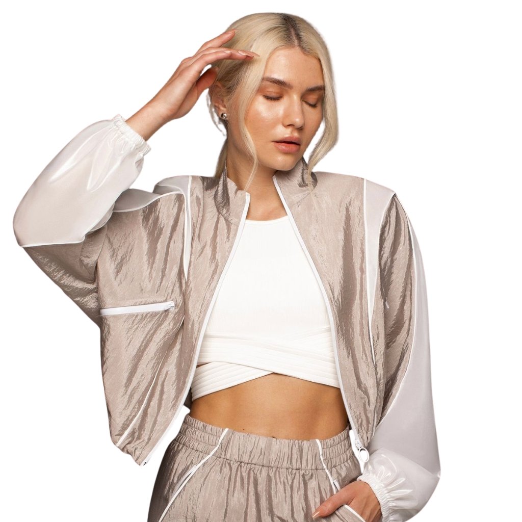 A woman with platinum blonde hair wearing a two-tone beige and white windbreaker over a white crop top, posing with one hand near her head against a white background.