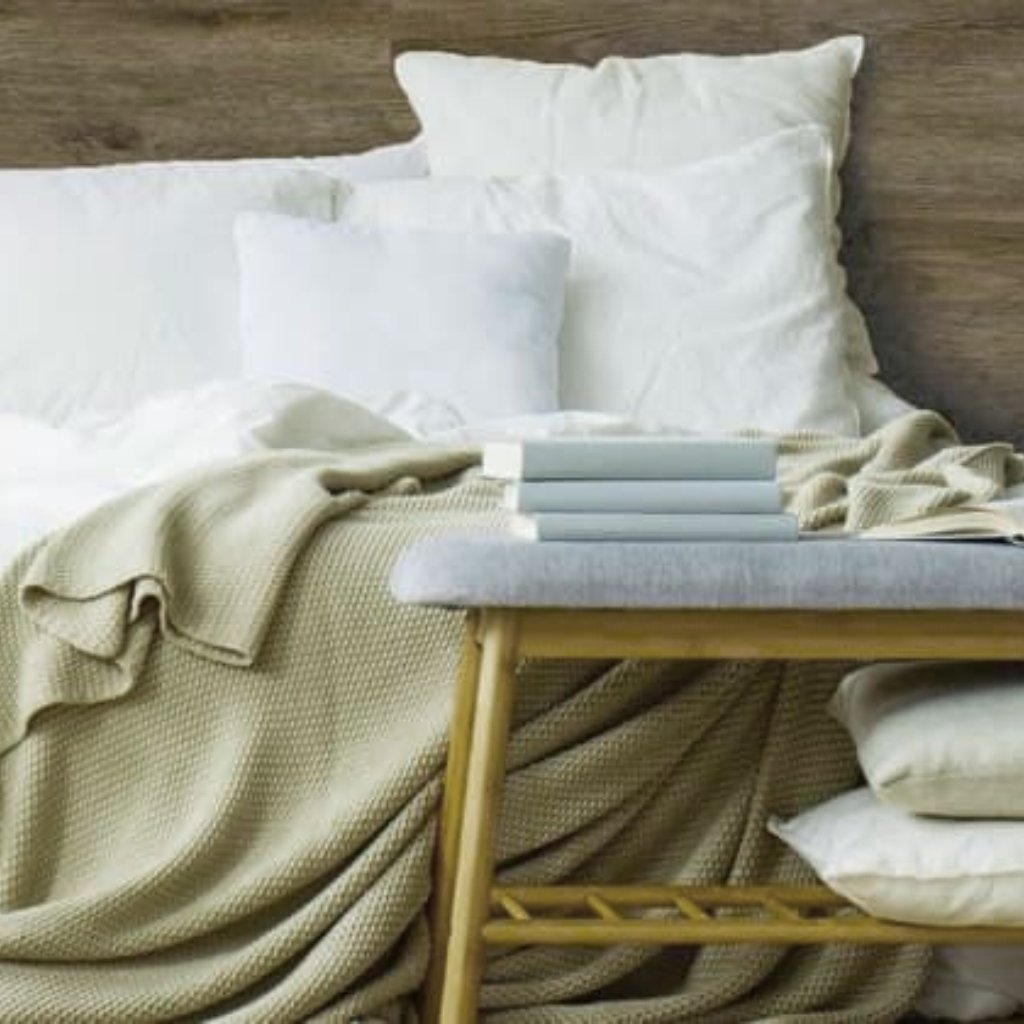 A cozy bedroom scene featuring a neatly made bed with white pillows and a beige knitted blanket. A light wooden bench at the foot of the bed holds a small stack of white books, with additional pillows and cushions nearby.