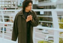 The Chesterfield Overcoat:Complete Guide & Modern Fashion A young person stands on a pedestrian bridge, wearing a long dark green coat and clasping their hands together as they look to the side, with city buildings and a railing in the background.