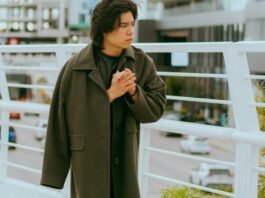 A young person stands on a pedestrian bridge, wearing a long dark green coat and clasping their hands together as they look to the side, with city buildings and a railing in the background.