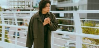 The Chesterfield Overcoat:Complete Guide & Modern Fashion A young person stands on a pedestrian bridge, wearing a long dark green coat and clasping their hands together as they look to the side, with city buildings and a railing in the background.