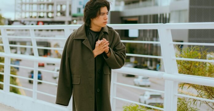 A young person stands on a pedestrian bridge, wearing a long dark green coat and clasping their hands together as they look to the side, with city buildings and a railing in the background.