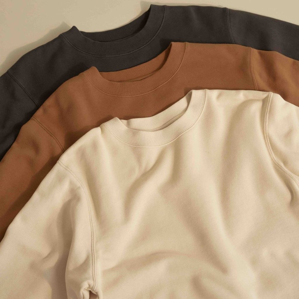 A stack of three crewneck sweatshirts in different colors (dark charcoal, warm brown, and off-white) arranged in a fan overlap.