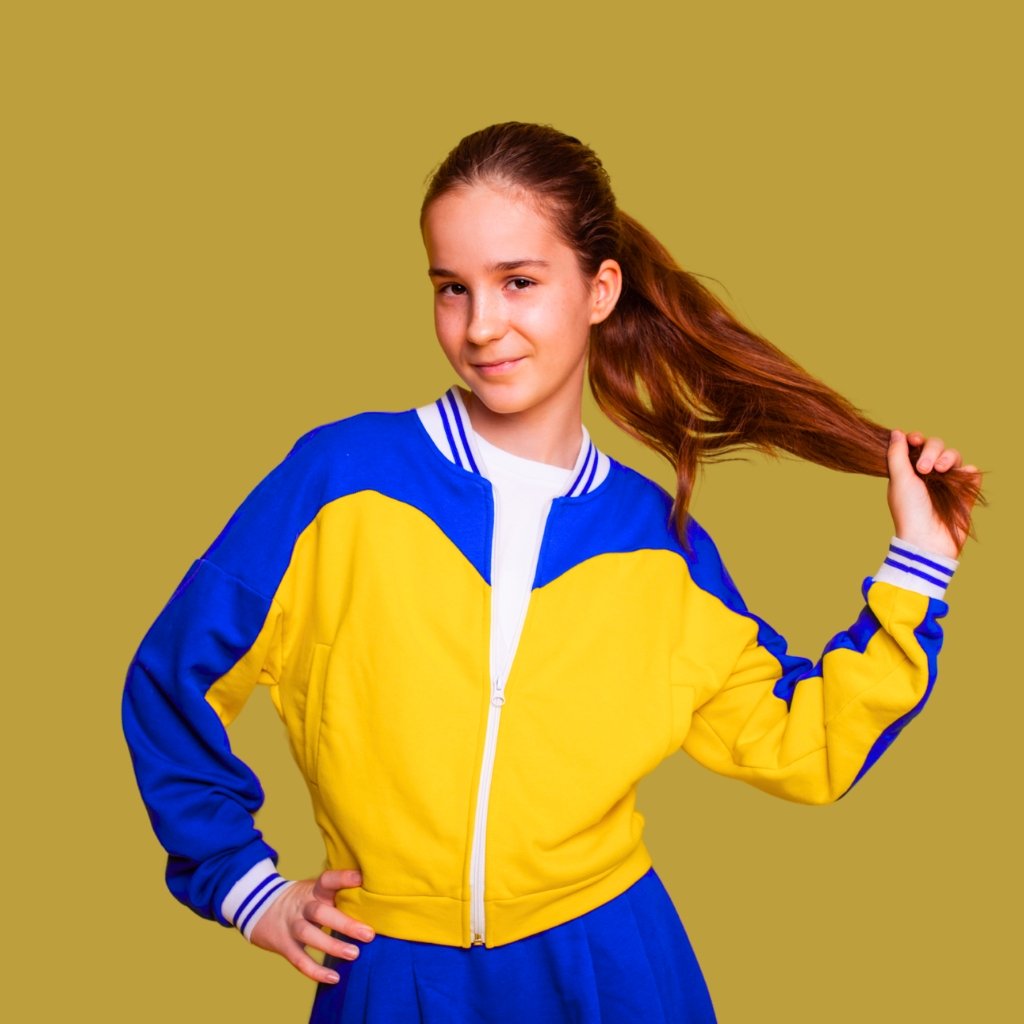 A young woman with long brown hair tied back, wearing a bright blue and yellow varsity jacket and smiling confidently while tugging her ponytail.