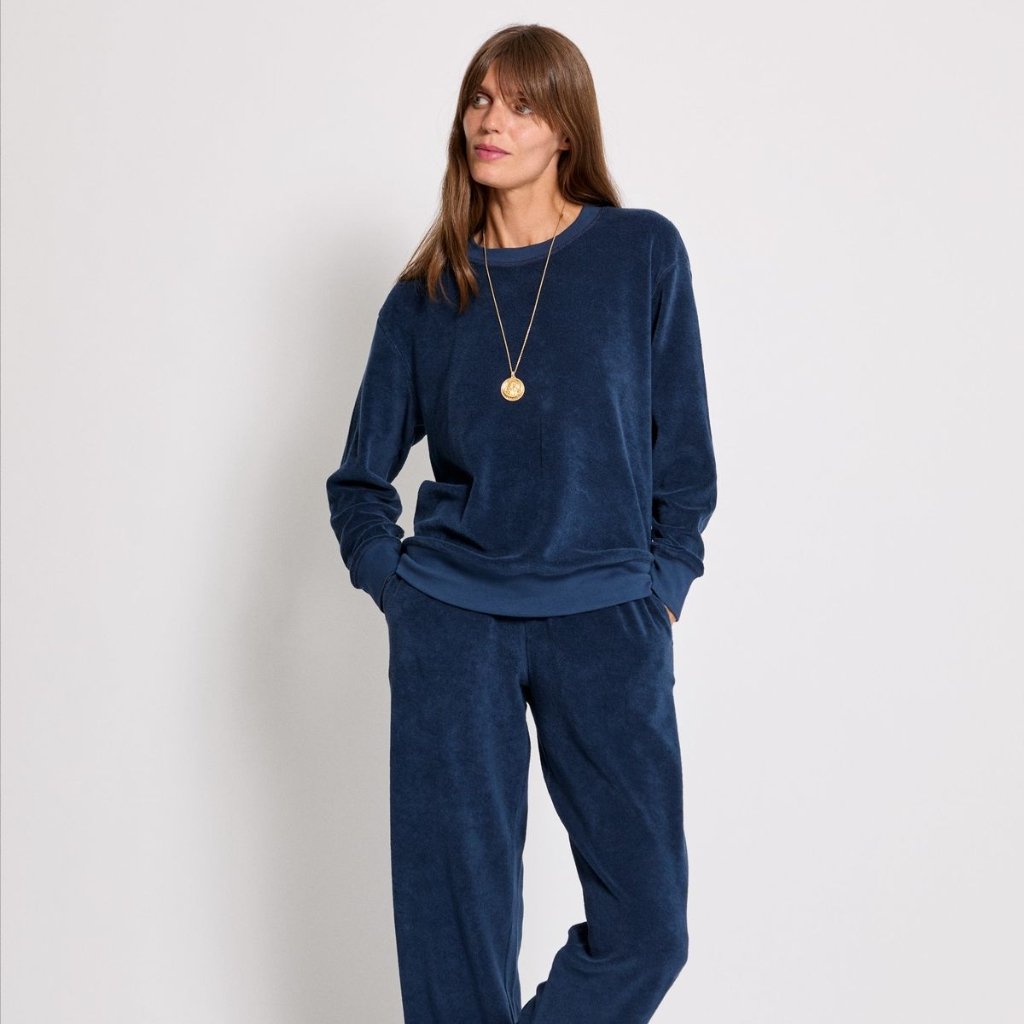 A woman wearing a matching navy blue velour tracksuit (crewneck sweatshirt and relaxed-fit pants) stands against a light background, hands in pockets, looking to the side. She wears a long pendant necklace.