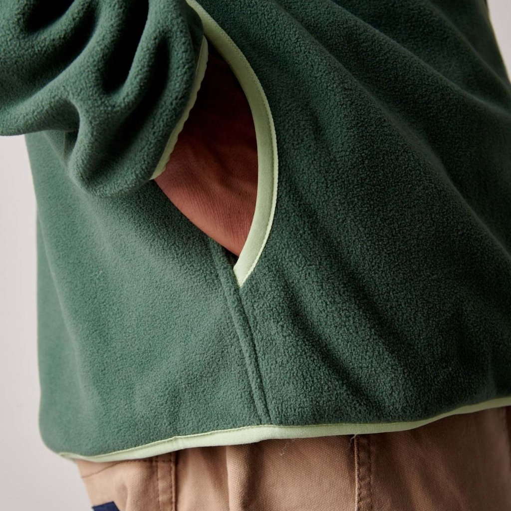 Close-up of a person wearing a dark green fleece jacket with light green piping at the pocket and hem, over brown pants.
