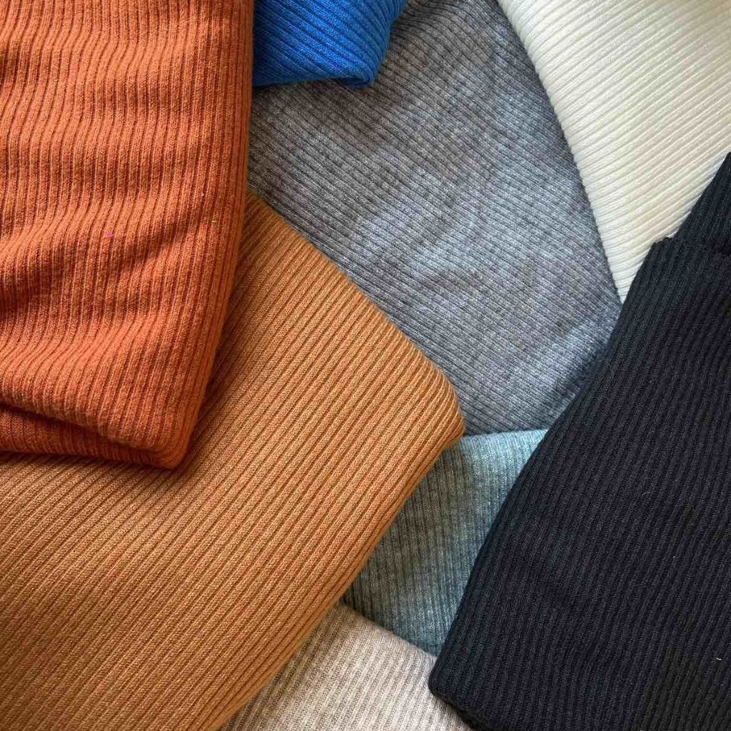 A close-up collage of folded ribbed knit fabrics in various colors (terracotta, rust, light beige, gray, blue, teal, cream, and black).

