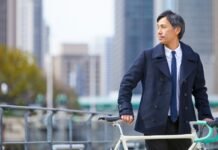 The Classic Peacoat A Complete Style Guide to Wearing A well-dressed man in a navy coat and tie stands beside a cream-colored bicycle on a city street with tall buildings in the background.