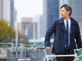 A well-dressed man in a navy coat and tie stands beside a cream-colored bicycle on a city street with tall buildings in the background.