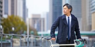 The Classic Peacoat A Complete Style Guide to Wearing A well-dressed man in a navy coat and tie stands beside a cream-colored bicycle on a city street with tall buildings in the background.