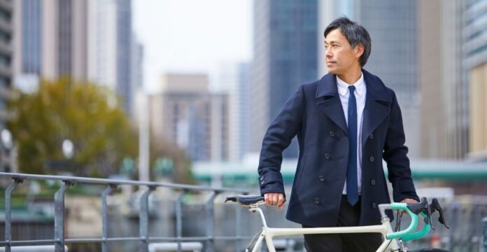 A well-dressed man in a navy coat and tie stands beside a cream-colored bicycle on a city street with tall buildings in the background.
