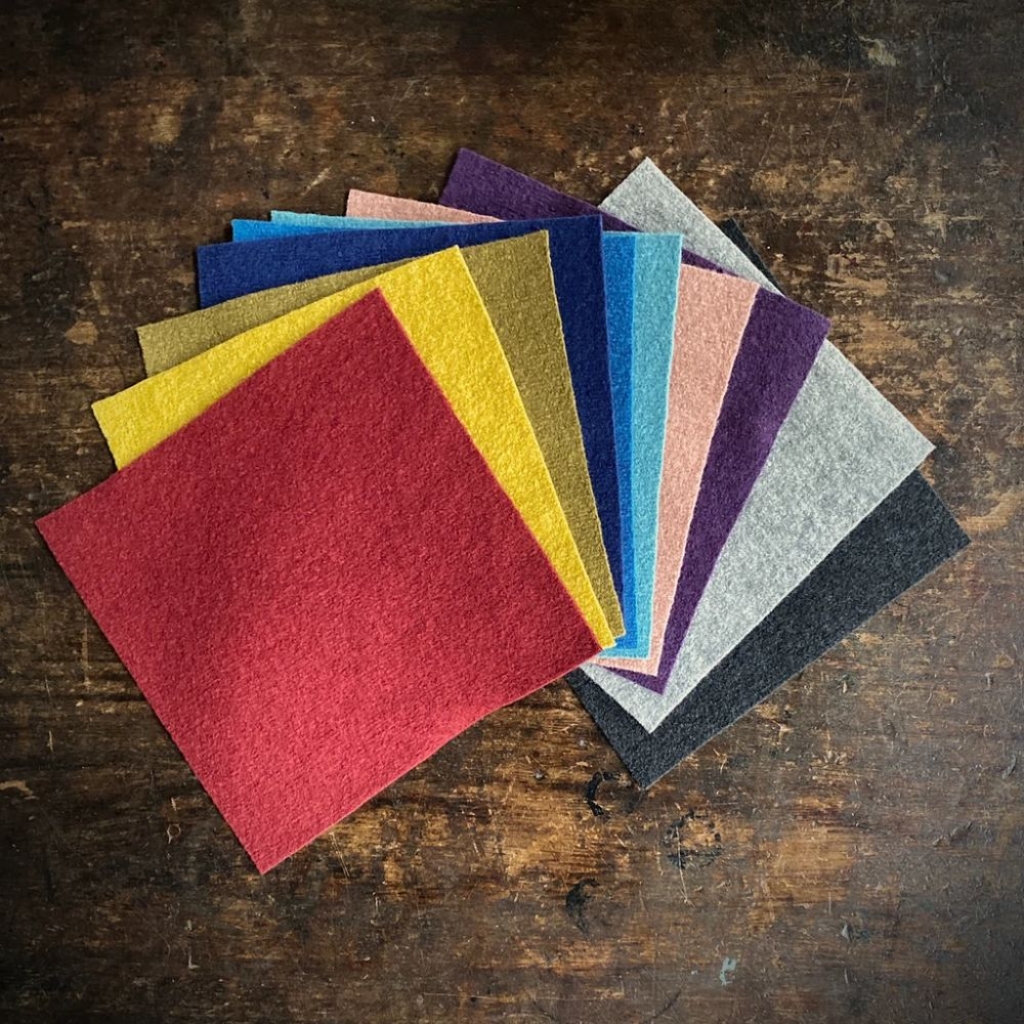 A fan of colorful felt squares arranged in a staggered arc on a dark wooden surface. The squares include red, yellow, gold, olive, blue, light blue, pink, purple, gray, and black tones.