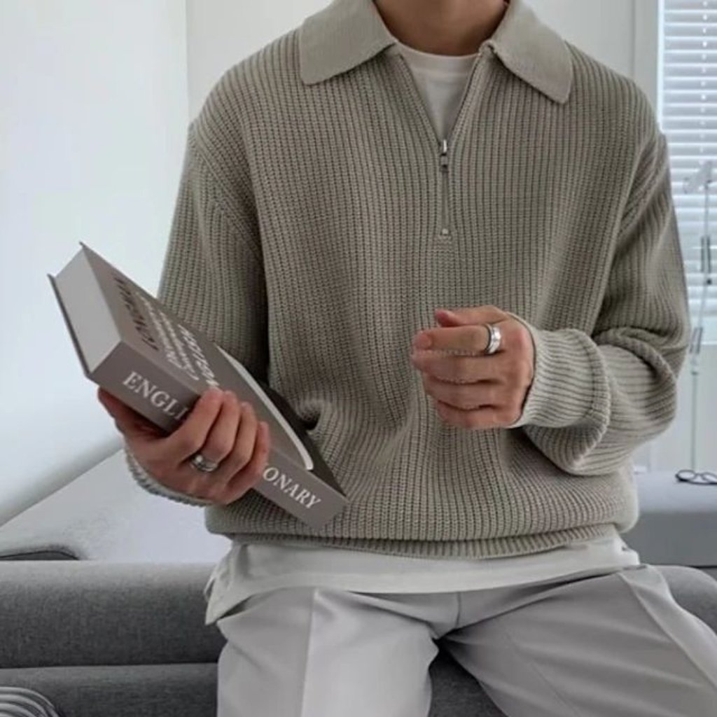 A person seated on a light-colored sofa, wearing a chunky knit beige pullover over a white shirt, holding a large dictionary in one hand and some rings on their fingers. The background shows a bright room with a window.

