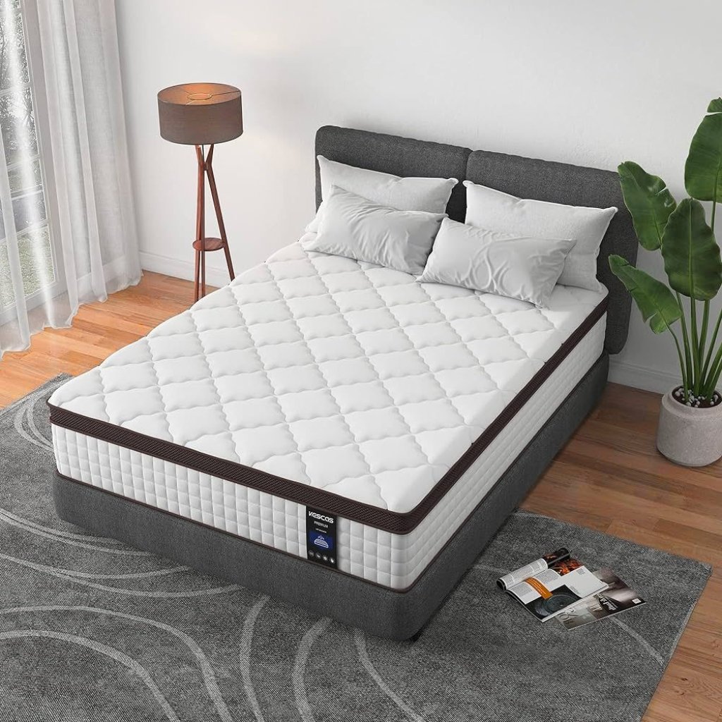 A neatly made bed with a white quilted mattress and dark brown trim sits on a gray upholstered bed frame in a bright, modern bedroom. A wooden floor lamp, sheer white curtains, a potted plant, and a gray area rug complete the space. A magazine lies open on the rug beside the bed.