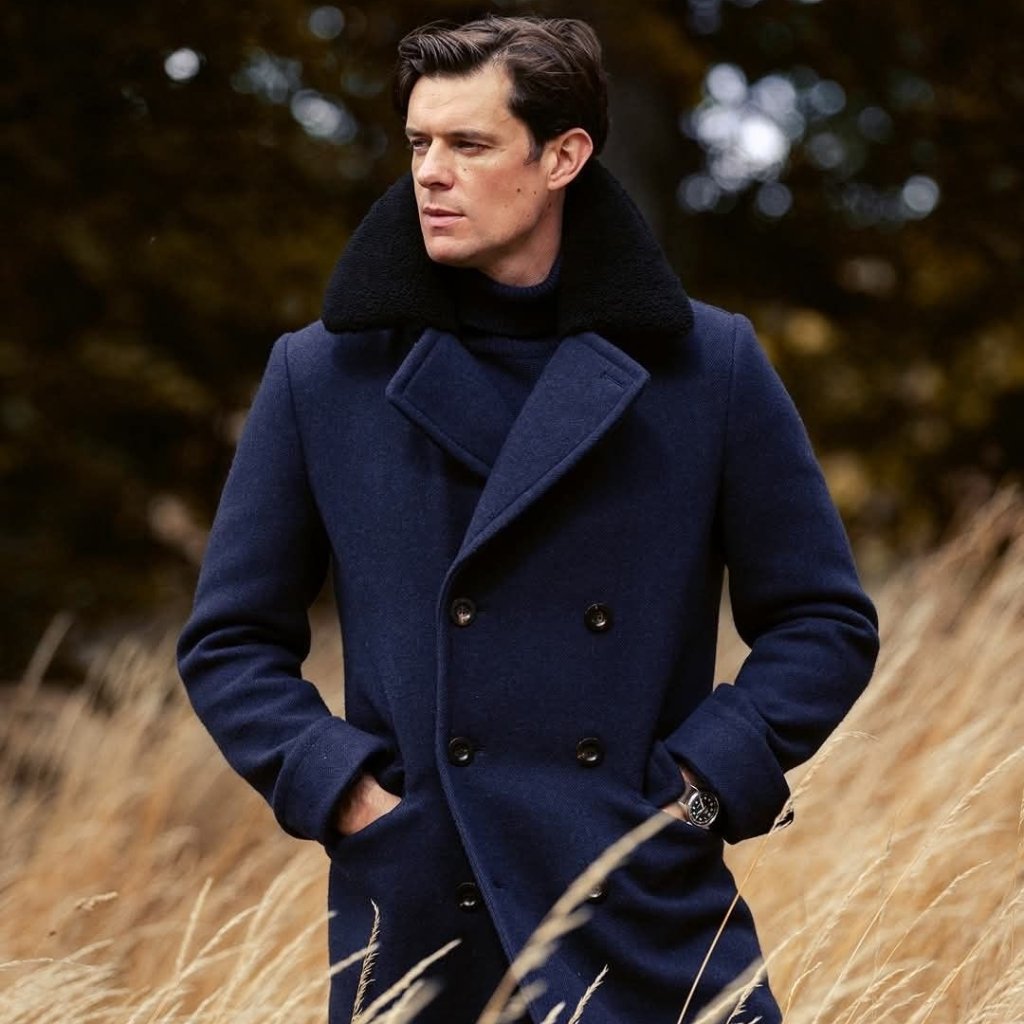 Topcoat Jackets Stylish Outerwear for a Sharp Seasonal Look 22 A man wearing a navy blue double-breasted wool coat with a black shearling collar stands outdoors in a field of tall, golden grass. He has hands in pockets and looks off to the side, with a dark watch on his left wrist.