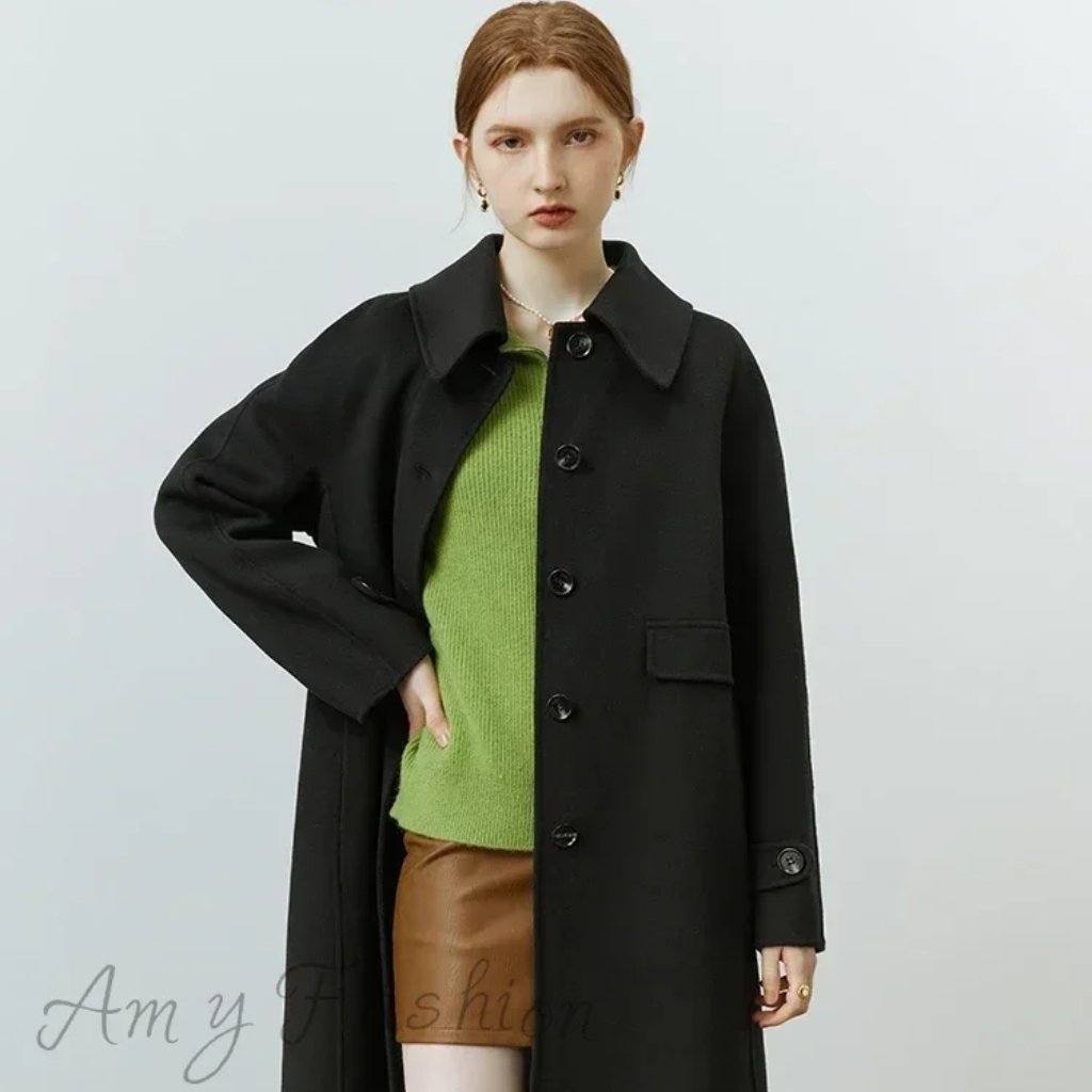 A fashion model wearing a long black coat over a bright green sweater and brown leather skirt, posing with one hand on her hip against a light background.