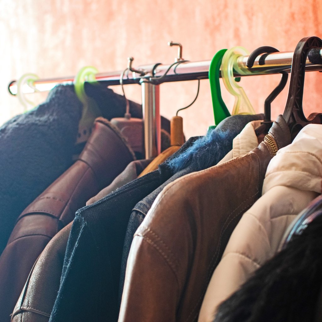 A row of jackets and coats hanging on a metal clothing rack with colorful hangers in green and black, against a warm, softly lit background . The image shows a clothing rack filled with various outerwear items, including leather, denim, and padded jackets, lined up on hangers. Bright green and clear hangers add pops of color, while the background features a soft, warm-toned wall. The scene evokes a sense of a boutique or wardrobe closet with well-organized attire ready for selection. UV Protection