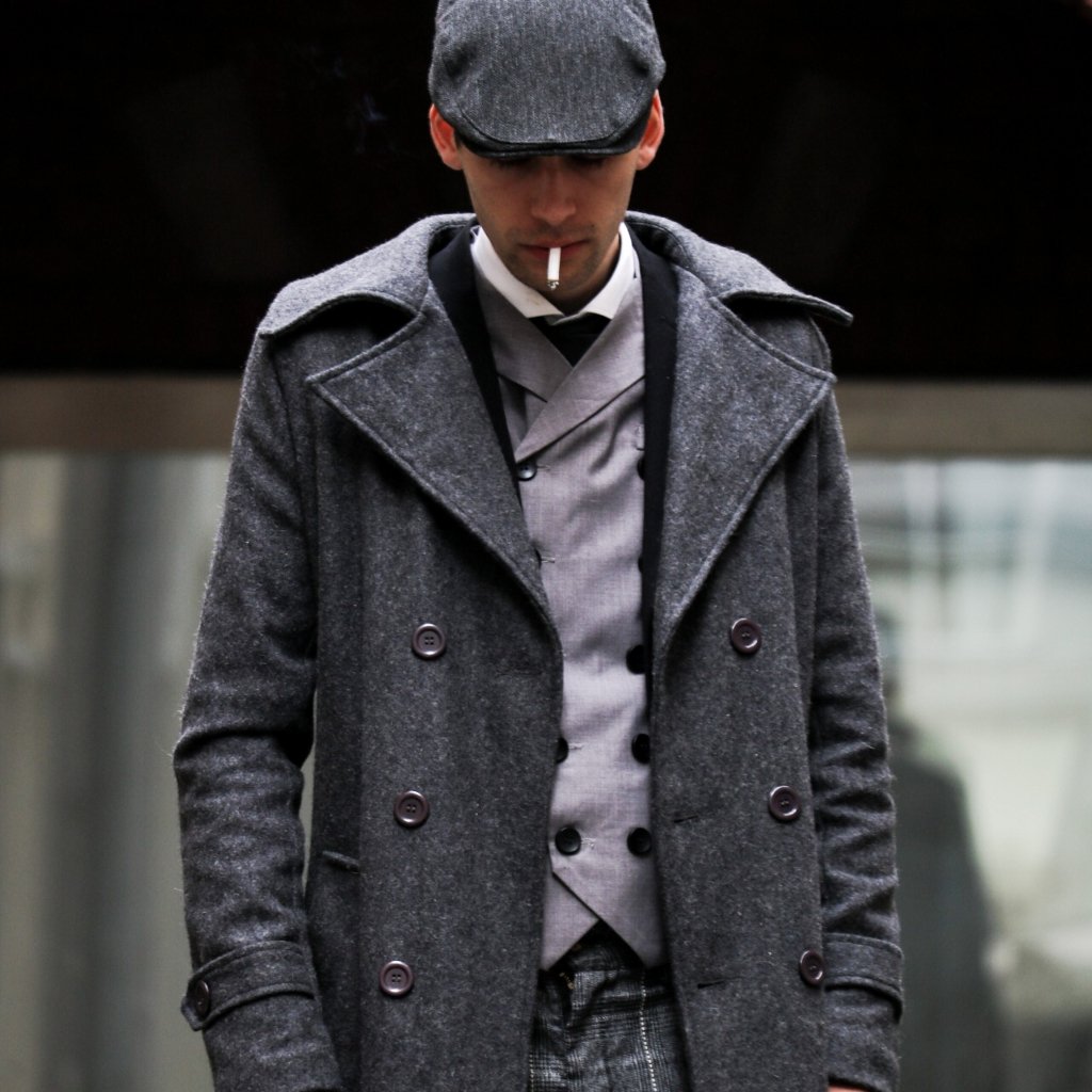 A man wearing a grey overcoat and flat cap looks downward while smoking a cigarette.