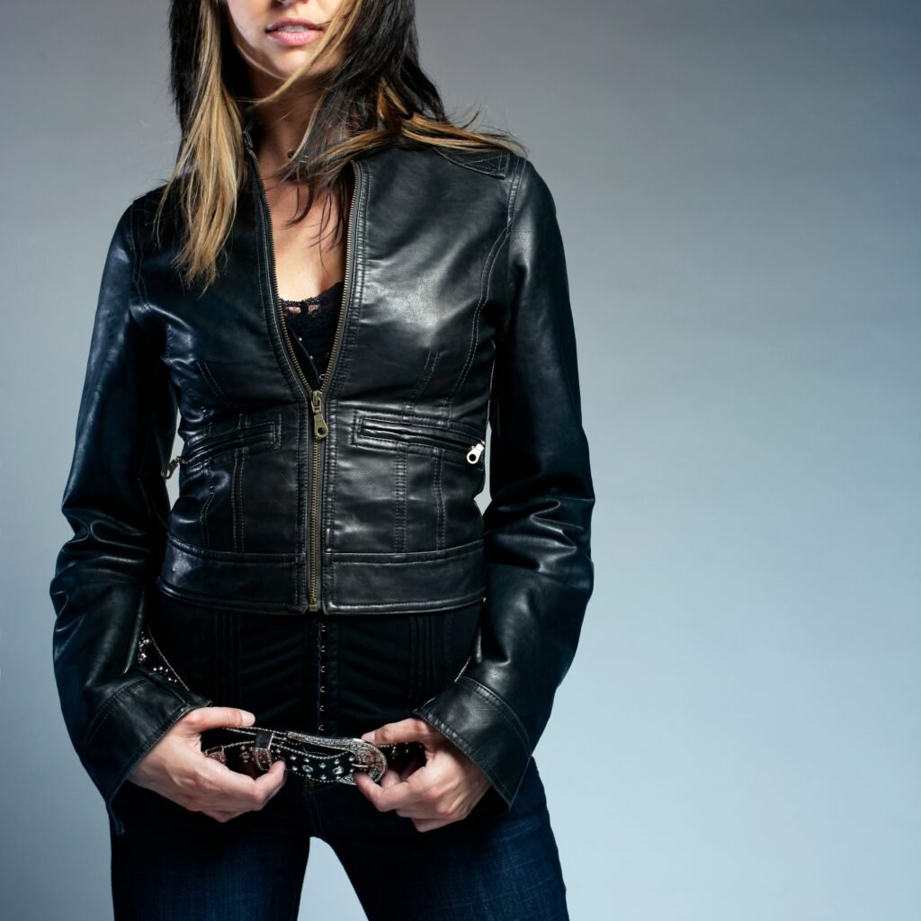 A person wearing a fitted black leather jacket with a front zipper, standing against a neutral, gray background. The jacket has seam details and zipped pockets; the person is holding a decorative belt with metal embellishments Field Leather  Jacket .