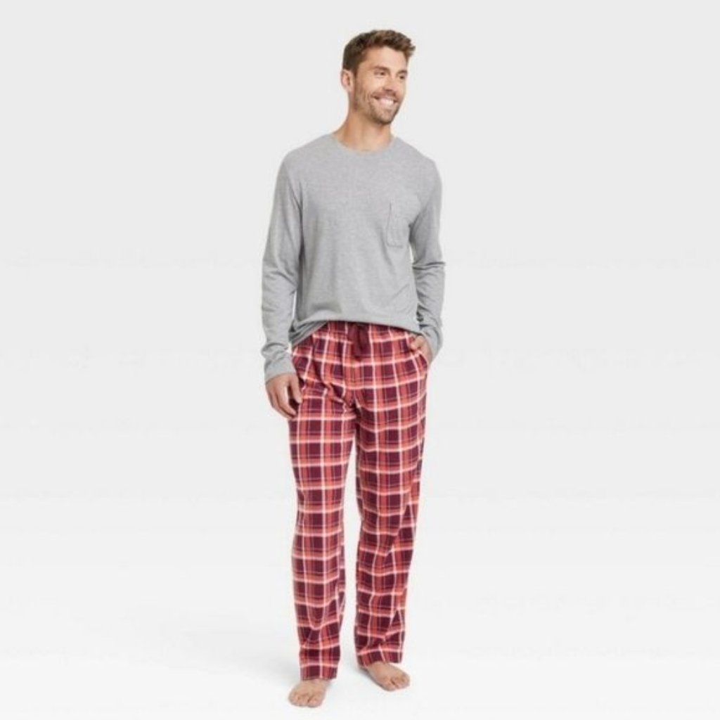 Man wearing a light gray long-sleeve shirt and red plaid pajama pants, standing barefoot with a relaxed smile