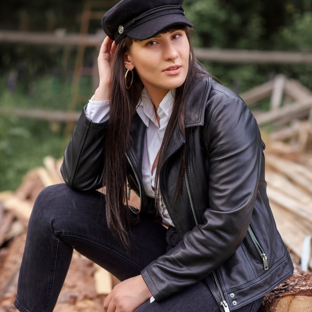 A woman sits outdoors on a stack of logs wearing a black leather field jacket, a white shirt, dark jeans, and a black cap. She has long dark hair and gold hoop earrings.Classic Field Leather Jacket