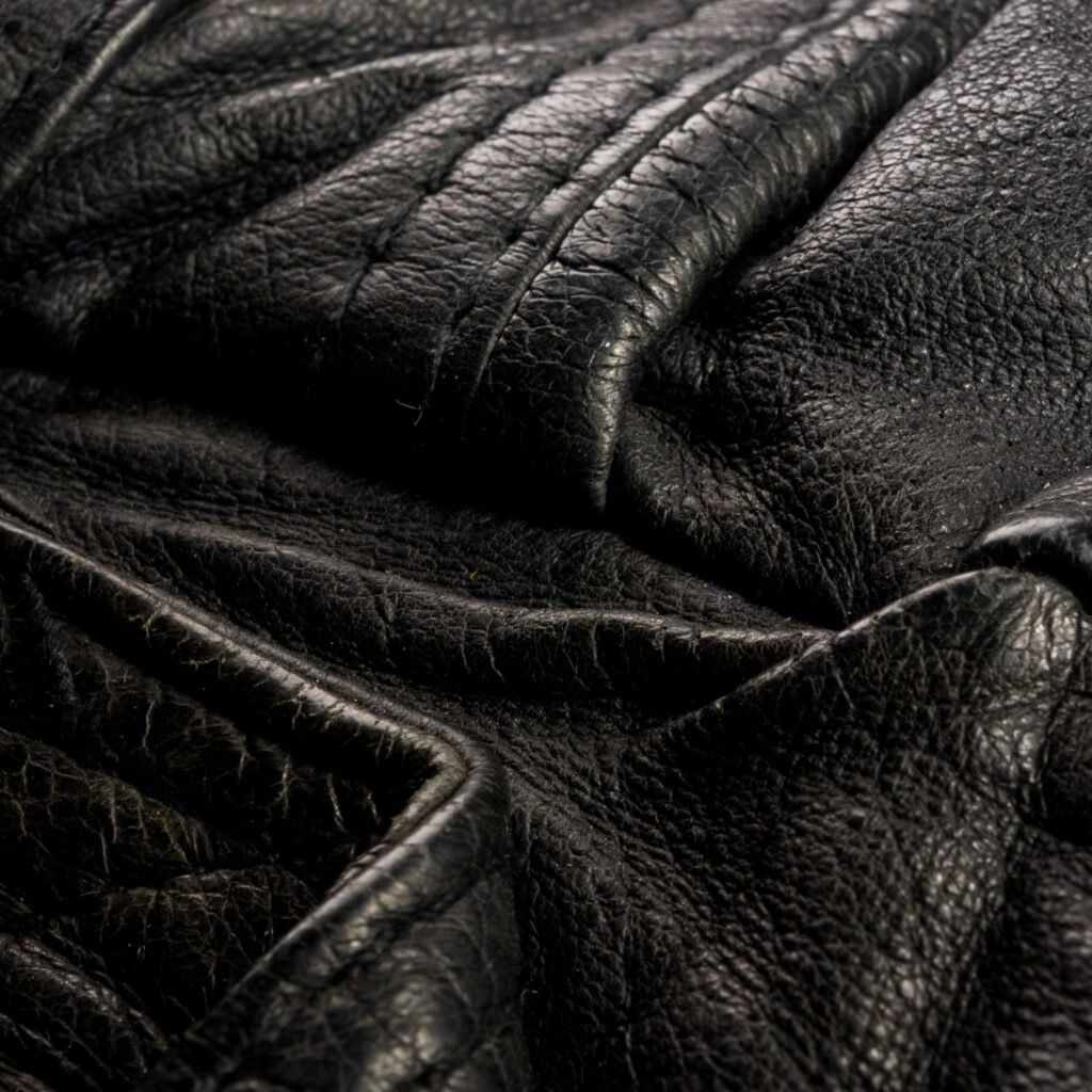 Close-up of black, textured leather with prominent stitching and natural creases, showcasing the supple grain and quilted or padded sections typical of a classic field jacket.