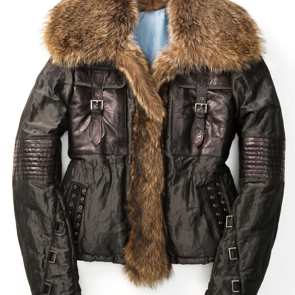 A black military-style field leather jacket with a thick fur-trimmed collar. The jacket features quilted shoulder patches, multiple buckle straps, and metal grommet details along the sides and sleeves. A blue garment is visible beneath.