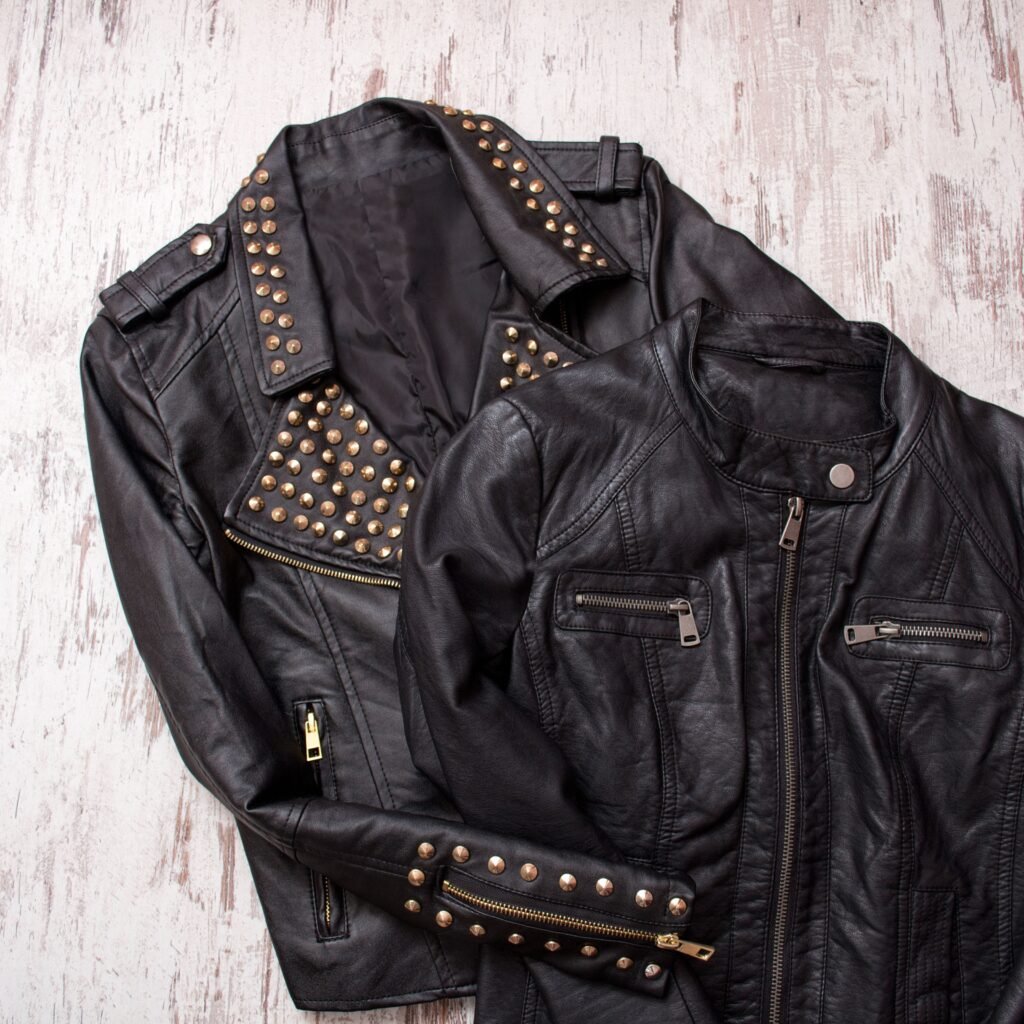 A black leather field jacket with brass studs along the collar, shoulders, and cuffs, laid on a light wooden surface.
