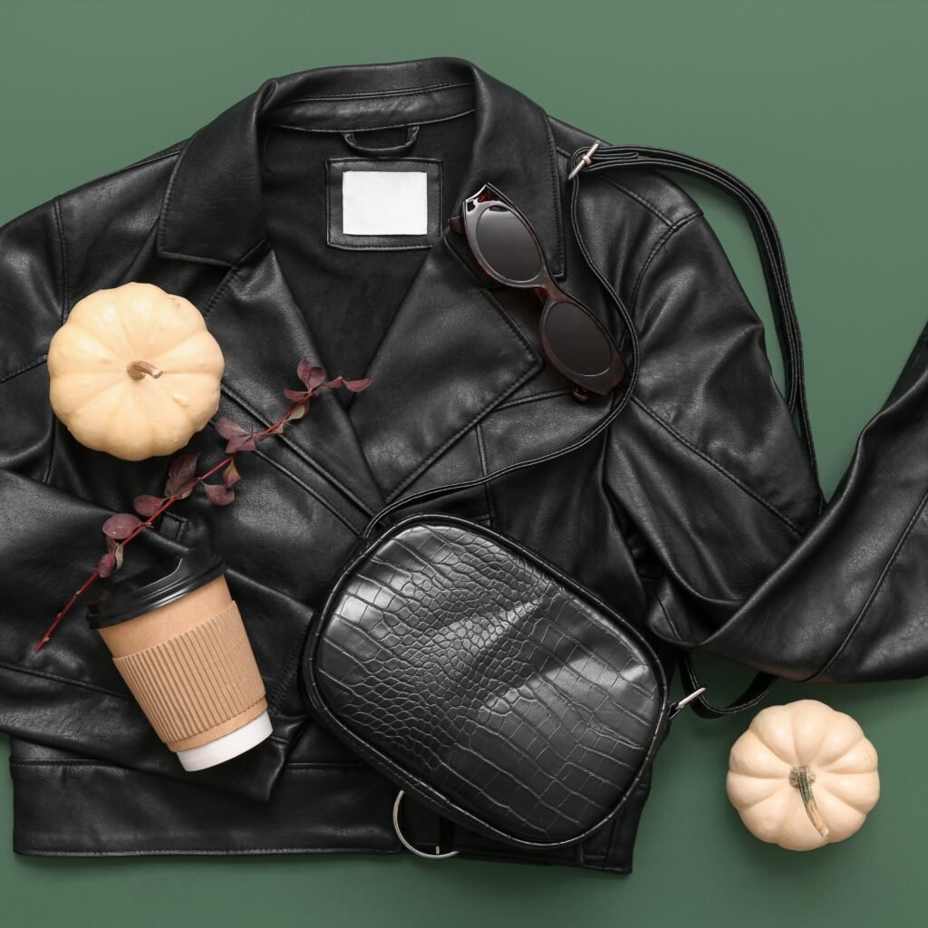 A black leather jacket laid flat with a small black crocodile-embossed shoulder bag, a pair of sunglasses, a white pumpkin, a disposable coffee cup with a brown sleeve, and a sprig of red leaves on a green background.