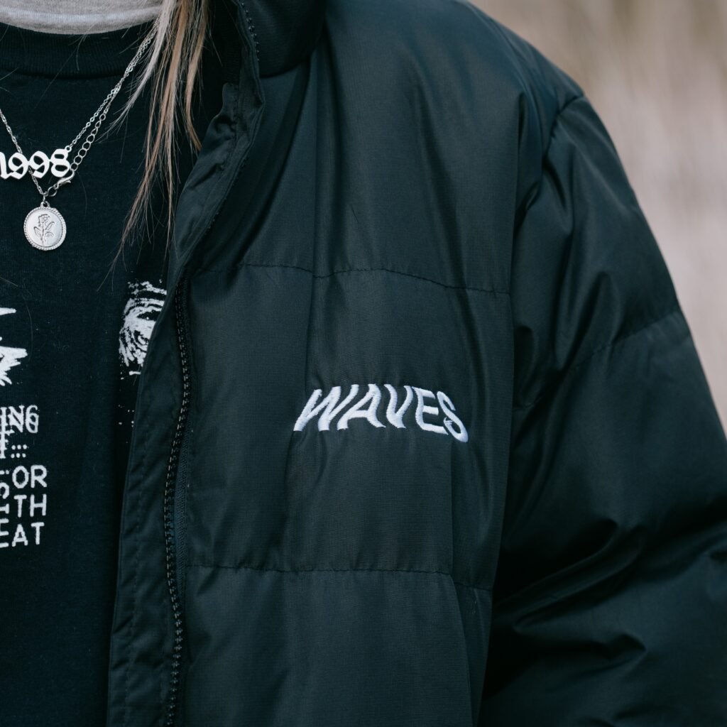 A person wearing a dark military-style field jacket with the word “WAVES” embroidered on the chest, paired with layered necklaces.