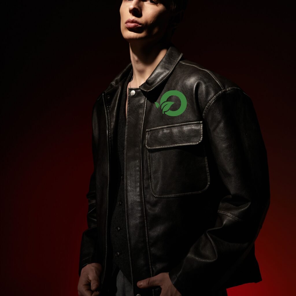 A person wearing a black leather field jacket with a green circular logo on the chest, standing against a dark red background.