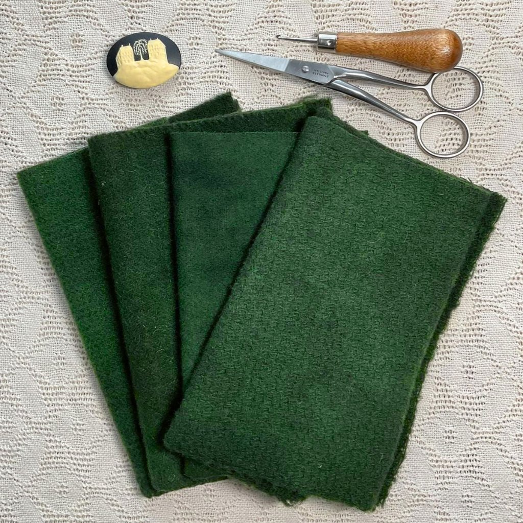 A collection of craft supplies on a lace tablecloth: four dark green felt sheets stacked in a fan, with a seam/rimmed pair of small scissors and a wooden-handled tool nearby; a small yellow-and-black pin or button sits at the top left.