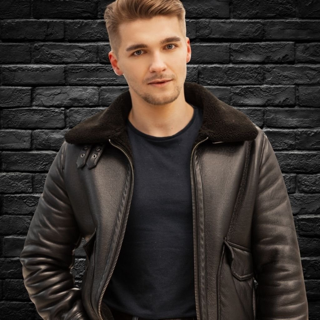 Modern Field Leather Jacket: Fresh Urban Look with Function 5 A man stands against a dark brick wall wearing a brown leather field jacket with a shearling collar and a black T-shirt.