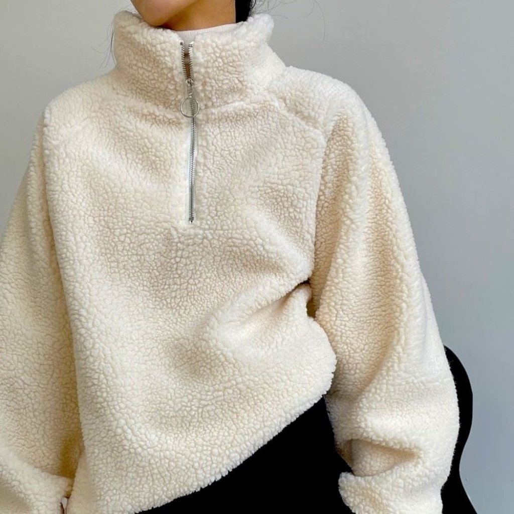 A close-up of a person wearing a cozy, oversized cream-colored sherpa fleece with a high collar and a front half-zip. The textured, fluffy material dominates the frame.