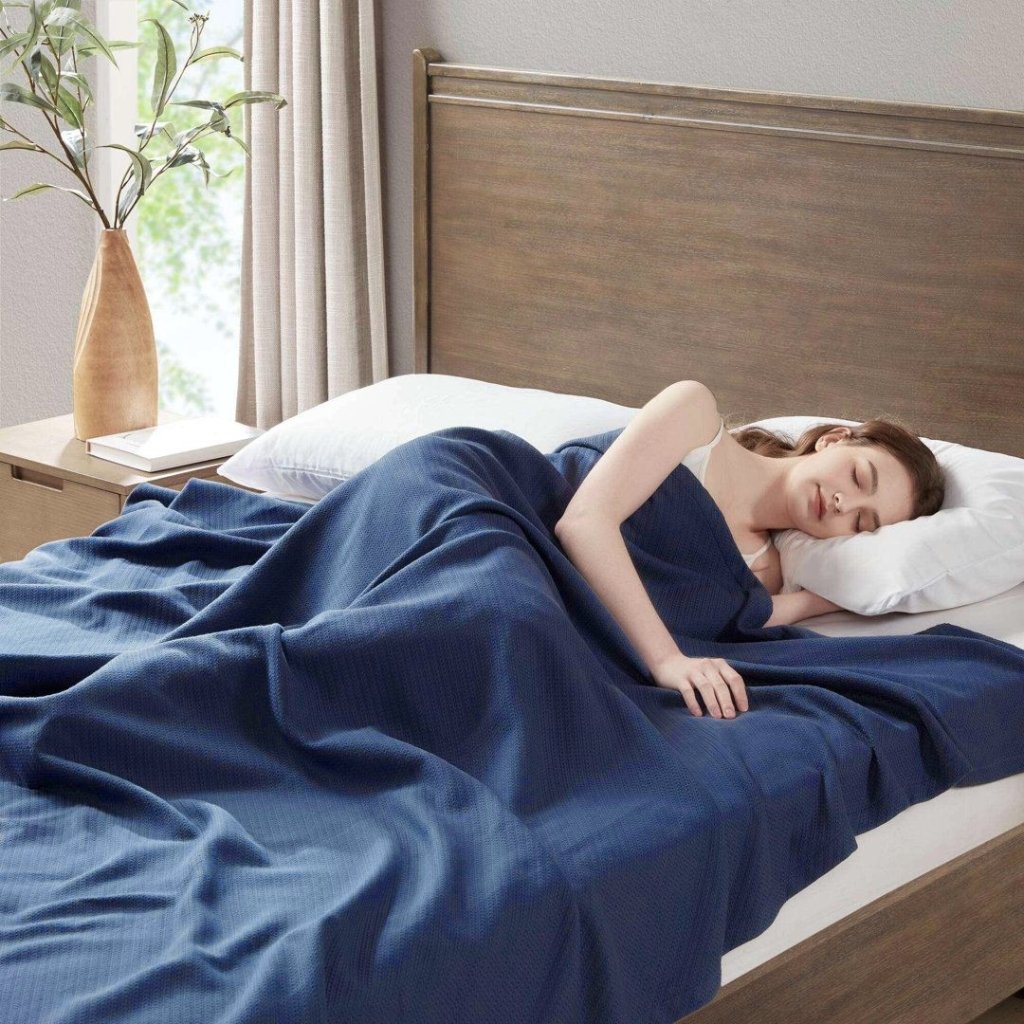 A woman sleeping on a bed, covered with a navy blue blanket, resting her head on a white pillow in a softly lit bedroom with a wooden headboard and a side table with a vase and book.