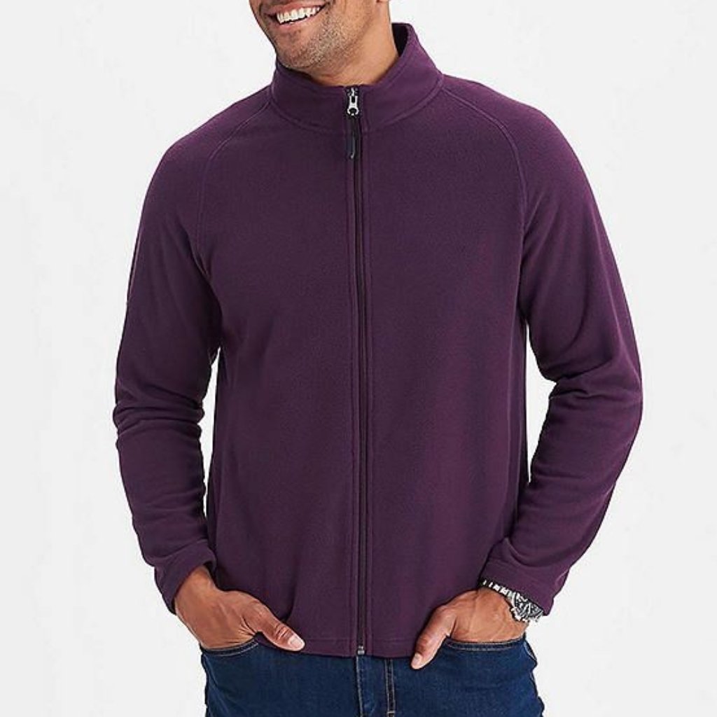 A man wearing a plain, zip-up purple fleece jacket with a high collar. He has his hands in his pockets and is smiling