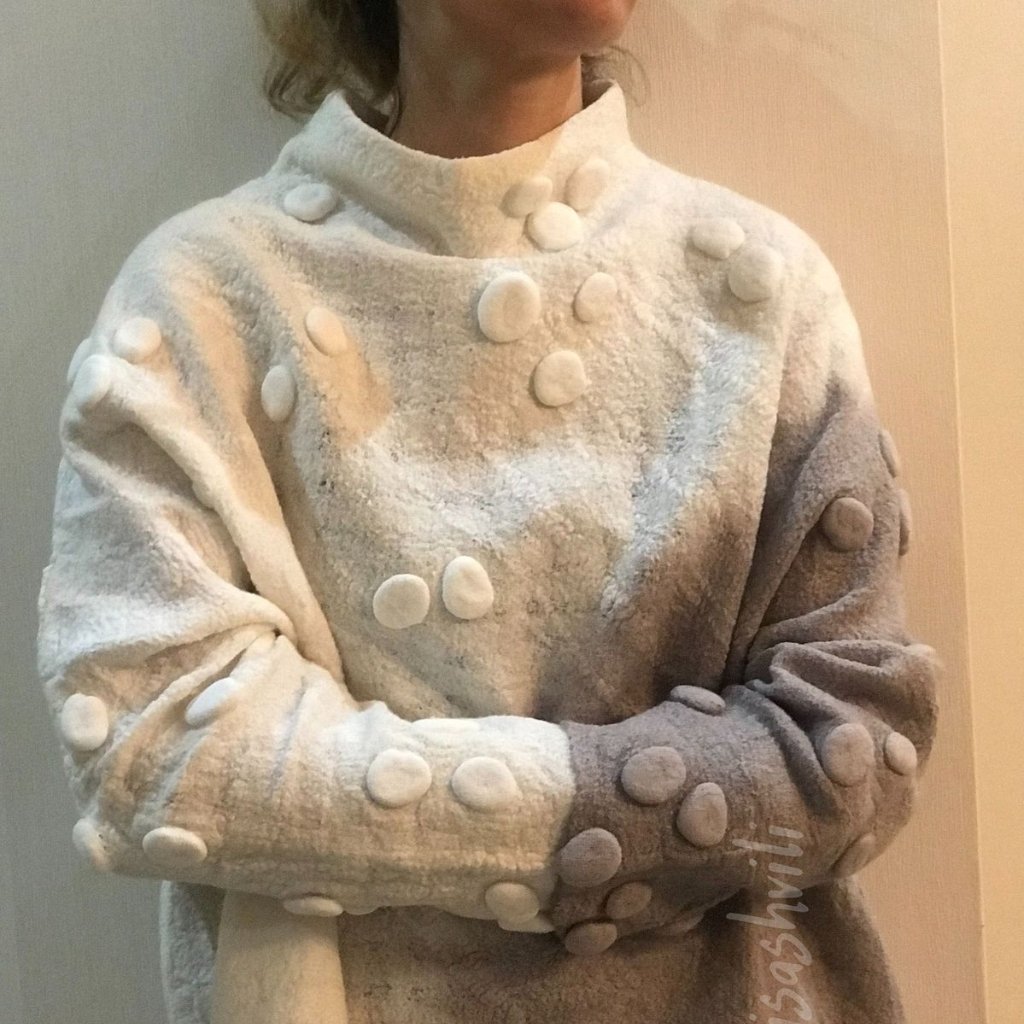 Felted Merino Fabric: Premium Softness for Cozy Elegance 9 A person wearing a fluffy, haute-collar sweater with large fabric buttons all over the front and sleeves, in a gradient light-to-dark gray. The person’s arms are crossed, and the photo is taken indoors against a neutral wall.