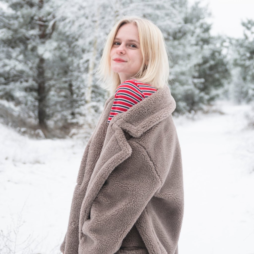 A young woman with short blonde hair stands in a snowy landscape, wearing a thick beige teddy coat over a red and white striped top, looking slightly over her shoulder with a soft smile.