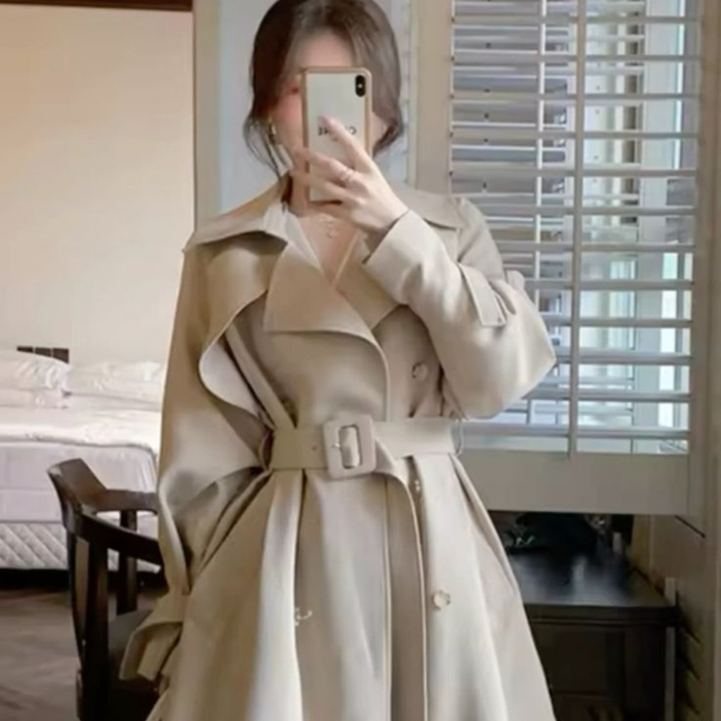 A person stands in a bedroom taking a mirror selfie with a smartphone. They wear a light beige trench coat with a wide belt, oversized lapels, and long sleeves. The background shows a bed, a chair, and a window with blinds.